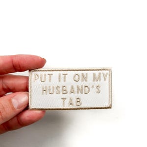Peut inclure: Patch en tissu blanc avec broderie beige qui dit "Put it on my husband's tab".