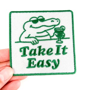 May include: Embroidered patch with a green border and a white background. The patch features a cartoon alligator wearing sunglasses and holding a drink. The text "Take It Easy" is embroidered in green.