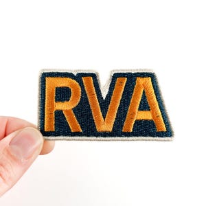 May include: Embroidered patch with the letters "RVA" in gold on a navy blue background. The patch is rectangular with a small cut-out at the top and a light border. The patch is held against a white backdrop.