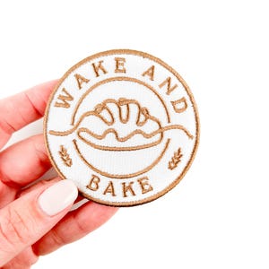 Può includere: Una toppa ricamata bianca con cuciture marroni. La toppa è rotonda e presenta un filone di pane, spighe di grano e il testo "Wake and Bake".