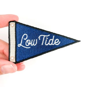 Pode incluir: Um patch em forma de flâmula azul marinho com as palavras "Low Tide" em escrita branca. O patch tem uma borda preta e uma borda branca texturizada com dois pequenos ilhós. O patch é segurado contra um fundo branco.