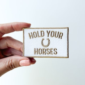 May include: White embroidered patch with gold thread. The patch says "Hold Your Horses" with a horseshoe symbol.