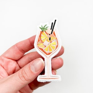 May include: A white embroidered iron-on patch of an orange cocktail with a slice of orange and a sprig of greenery in a stemmed glass.