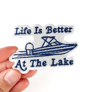 May include: Embroidered patch with a white background and a blue boat design. The text "Life Is Better At The Lake" is embroidered in blue. The patch is held by a hand.