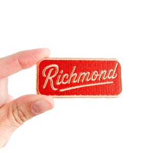 May include: A red embroidered patch with the name "Richmond" in cream cursive lettering. The patch has a gold border and is held up by a hand against a white backdrop. This patch could be for a jacket or bag, and is rectangular in shape.