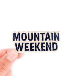 May include: A rectangular patch with the words "MOUNTAIN WEEKEND" embroidered in navy blue on a cream-colored background. The patch is held up against a white background.
