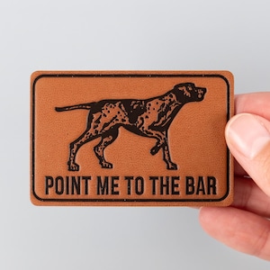 Könnte beinhalten: Ein rechteckiger, hellbrauner Lederaufnäher mit einer schwarzen Illustration eines Hundes und dem Schriftzug "POINT ME TO THE BAR". Der Hund ist ein Vorstehhund, mit geflecktem Fell. Der Aufnäher wird von einer Hand gehalten.