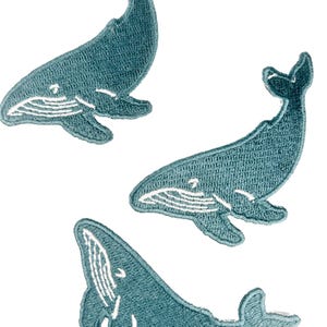 May include: Three embroidered patches in the shape of whales. The patches are teal blue with white detailing, including the whale's belly and facial features. The patches are designed to be sewn onto fabric.