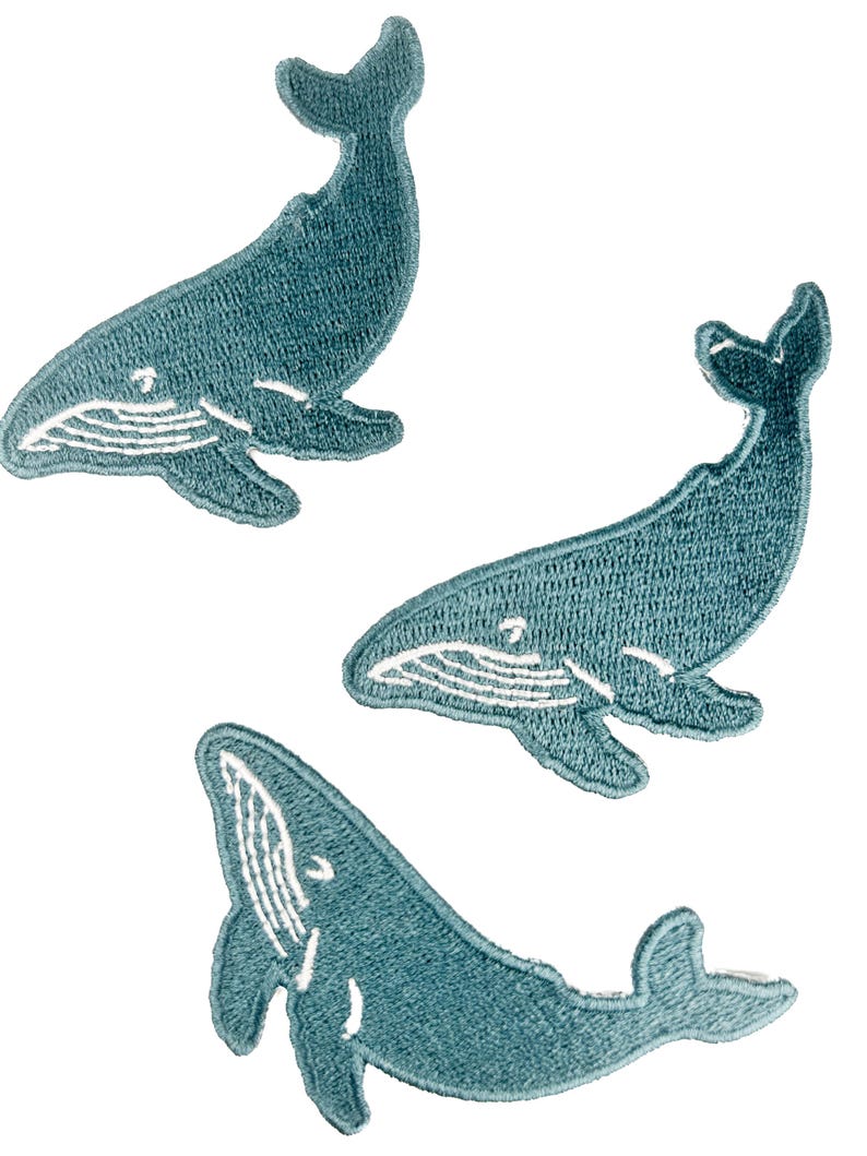 May include: Three embroidered patches in the shape of whales. The patches are teal blue with white detailing, including the whale's belly and facial features. The patches are designed to be sewn onto fabric.