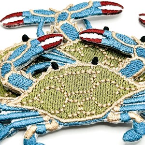 May include: A collection of embroidered crab patches featuring a light green shell with tan stitching details, bright blue legs, and claws tipped with deep red. The crab designs are layered, showcasing the intricate thread work and texture.