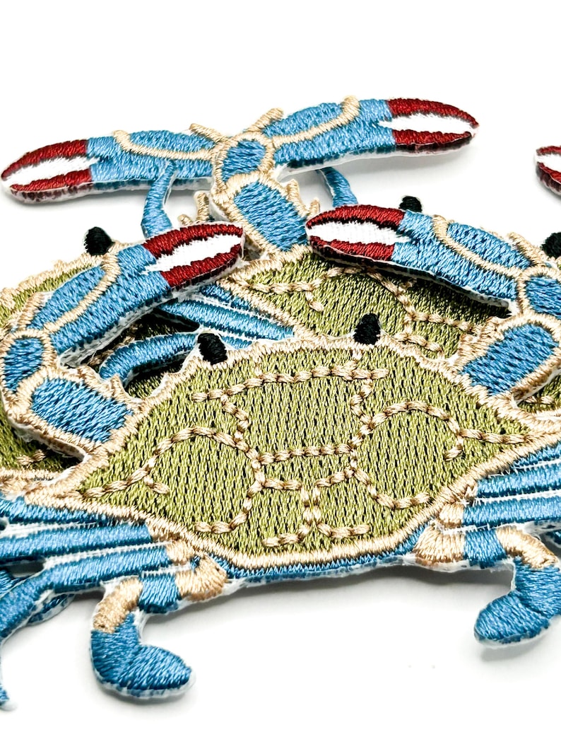 May include: A collection of embroidered crab patches featuring a light green shell with tan stitching details, bright blue legs, and claws tipped with deep red. The crab designs are layered, showcasing the intricate thread work and texture.