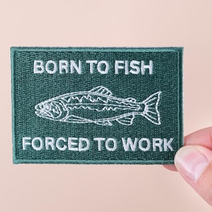 Puede incluir: Un parche rectangular bordado en verde oscuro con texto blanco y una ilustración de un pez. El texto dice "BORN TO FISH" encima de un contorno de pez y "FORCED TO WORK" debajo. El parche está sostenido por una mano.