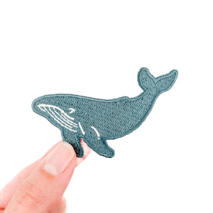 Puede incluir: Un parche bordado de ballena de color verde azulado con detalles blancos. El parche presenta un diseño de ballena con un cuerpo curvo y una cola detallada. El parche se sostiene sobre un fondo blanco.