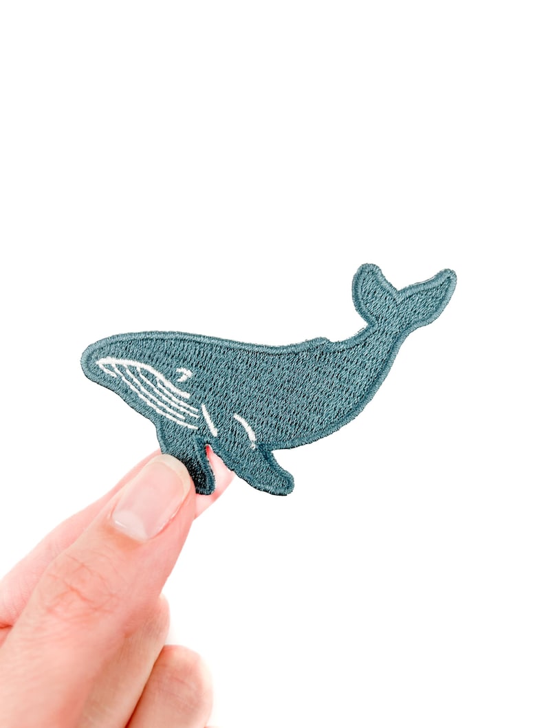 May include: A teal embroidered whale patch with white detailing. The patch features a whale design with a curved body and a detailed tail. The patch is held up against a white background.