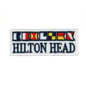 May include: Rectangular embroidered patch with the text "HILTON HEAD" in navy blue block letters. Above the text are colorful nautical signal flags. The patch has a white border and is likely for a jacket or bag.