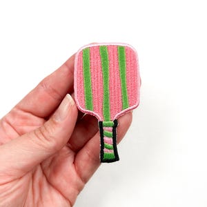 May include: An embroidered patch shaped like a pickleball paddle, with pink and green stripes.