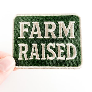 Op de afbeelding: Een rechthoekige, geborduurde patch met de woorden "FARM RAISED" in lichtgrijze letters. De patch heeft een donkergroene achtergrond en een witte rand. De patch wordt door een hand tegen een witte achtergrond gehouden.