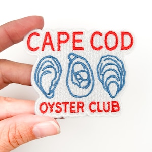 Peut inclure: Écusson brodé avec le texte "CAPE COD OYSTER CLUB" en rouge, au-dessus de trois dessins de coquilles d'huîtres bleues. L'écusson est blanc avec un bord rouge et est tenu par une main.