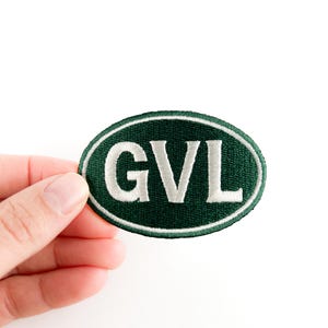 Può includere: Toppa ovale con le lettere bianche "GVL" ricamate su sfondo verde scuro. La toppa ha un bordo bianco. La toppa è tenuta su uno sfondo bianco.