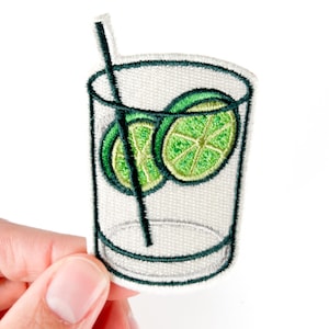 May include: Embroidered patch featuring a cocktail glass with two lime slices and a black straw. The glass is outlined in black, with green and white stitching. The patch is held against a white background.