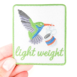 May include: Embroidered patch featuring a hummingbird in flight, with green, blue, and purple details, carrying a small object. The patch has a white background with the words "light weight" in green script, framed by a green border.