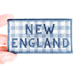 May include: A rectangular patch with the words "NEW ENGLAND" embroidered in navy blue thread. The patch has a blue and white gingham pattern background and a navy blue border.