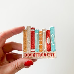 Op de afbeelding: Een geborduurde strijk-op patch met een stapel kleurrijke boeken met de tekst "BOOKTROVERT" eronder.