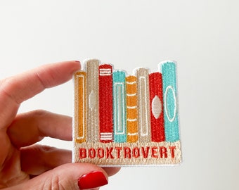 Pode incluir: Um remendo bordado para passar a ferro com uma pilha de livros coloridos com o texto "BOOKTROVERT" abaixo.