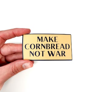 Puede incluir: Una pegatina amarilla con texto negro que dice "Make Cornbread Not War".