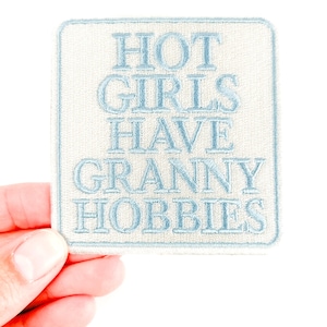 May include: A square patch with the text "HOT GIRLS HAVE GRANNY HOBBIES" embroidered in light blue thread on a cream-colored background. The patch is held by a hand against a white background.