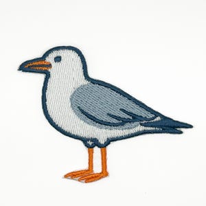 May include: An embroidered patch featuring a seagull in profile. The bird has a white head and chest, light blue wings with dark blue outlines, a dark blue beak with an orange tip, and bright orange legs and webbed feet on a white background.