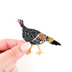 Puede incluir: Un parche de pavo bordado en negro, naranja y marrón con una barba roja. El pavo está de pie sobre dos patas naranjas con tres dedos cada una.