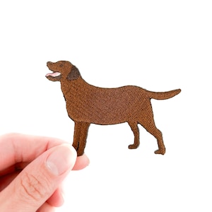 May include: A brown embroidered patch in the shape of a Labrador Retriever dog. The dog is standing with its mouth open, showing a pink tongue. The patch is held against a white background.