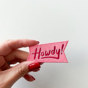 Puede incluir: Un parche bordado rosa con la palabra "Howdy!" en cursiva. El parche tiene forma de bandera con un fondo rosa claro y letras rosa oscuro. El parche está sostenido por una mano con esmalte de uñas rojo.