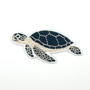 以下が含まれることがあります： ウミガメの形をした刺繍入りのアイロン接着ワッペン。カメの甲羅は濃いネイビーブルーで白いステッチが施されており、腹部はライトベージュ、頭部とヒレはネイビーブルーと白の糸で模様が描かれています。