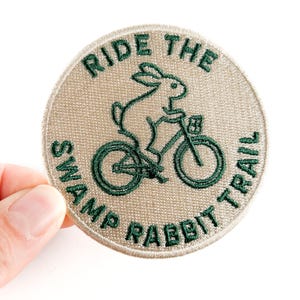 Może przedstawiać: Okrągła, haftowana naszywka z napisem "RIDE THE SWAMP RABBIT TRAIL" w kolorze zielonym. Projekt przedstawia królika jadącego na rowerze, również w kolorze zielonym, na beżowym tle. Naszywka ma obszytą krawędź.