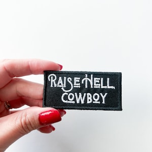 Op de afbeelding: Zwarte geborduurde patch met witte tekst die "Raise Hell Cowboy" luidt.