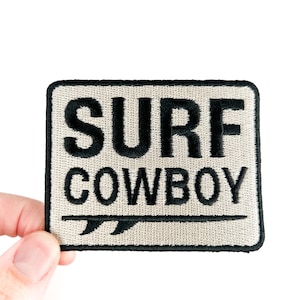 May include: Embroidered patch with the words "SURF COWBOY" in black block letters on a beige background. A black surfboard graphic is below the word "COWBOY". The patch has a black border.