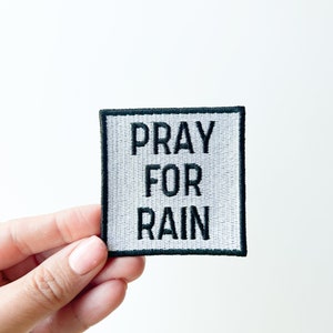 May include: A white fabric patch with black embroidered text that reads "Pray for Rain". The patch has a black border.