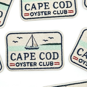 Może przedstawiać: Prostokątne haftowane naszywki przedstawiające żaglówkę na jasnym, zielono-kremowym tle. Napis CAPE COD OYSTER CLUB wykonano ciemnoniebieską nicią, z dwiema małymi czerwonymi poziomymi liniami po obu stronach słowa OYSTER.