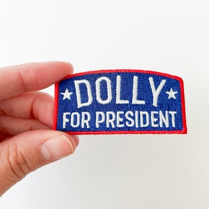 Puede incluir: Un parche bordado azul y rojo con texto blanco que dice "*DOLLY* FOR PRESIDENT" con una estrella blanca a cada lado de la palabra "DOLLY".