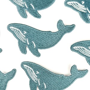 May include: Embroidered patches featuring a teal whale design with white detailing. The patches are in a repeating pattern on a white background. The whale patches are a cute and whimsical design.