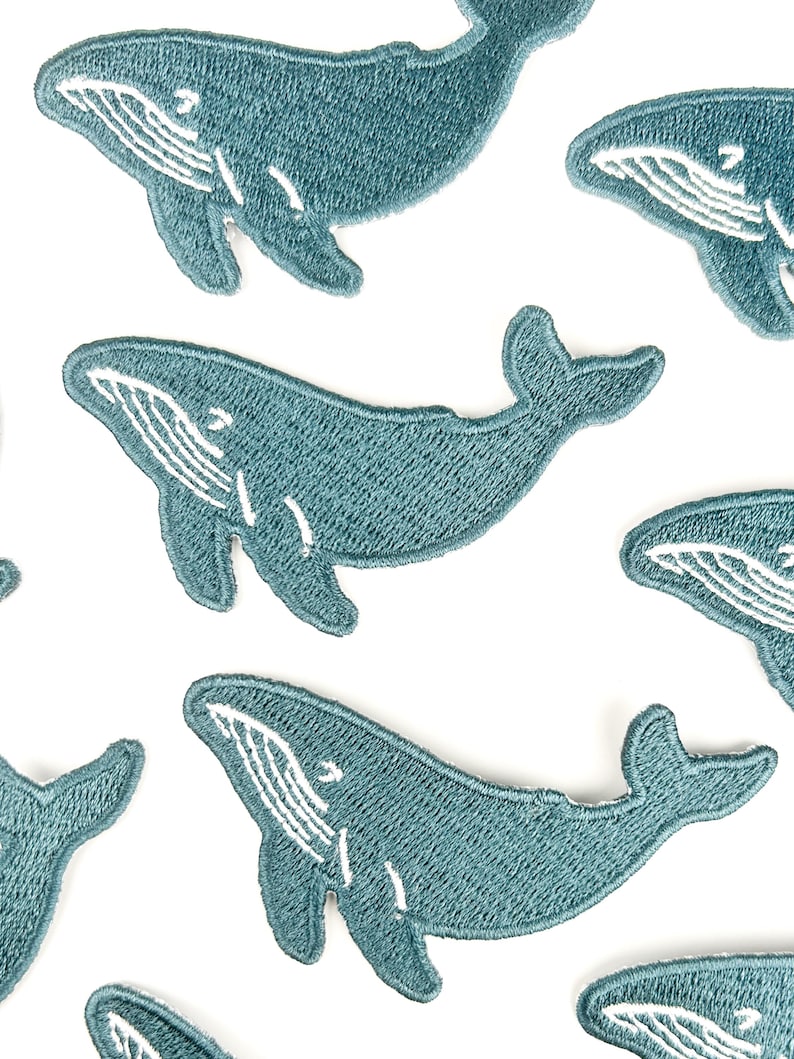 May include: Embroidered patches featuring a teal whale design with white detailing. The patches are in a repeating pattern on a white background. The whale patches are a cute and whimsical design.