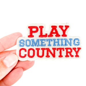 Può includere: Toppa ricamata con le parole "PLAY SOMETHING COUNTRY" in lettere rosse e blu. La toppa ha un bordo bianco ed è tenuta su uno sfondo bianco.