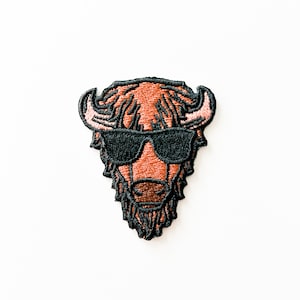 May include: Embroidered patch of a brown buffalo wearing black sunglasses. The buffalo has a black beard and horns.