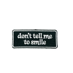 May include: A black rectangular patch with white text that reads "don't tell me to smile". The patch has a white border and is likely an iron-on or sew-on patch for clothing or accessories.