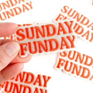 Puede incluir: Parches bordados con las palabras "SUNDAY FUNDAY" en hilo naranja sobre un fondo blanco. Los parches son rectangulares y tienen un borde cosido. Varios parches est&aacute;n dispersos sobre una superficie blanca.