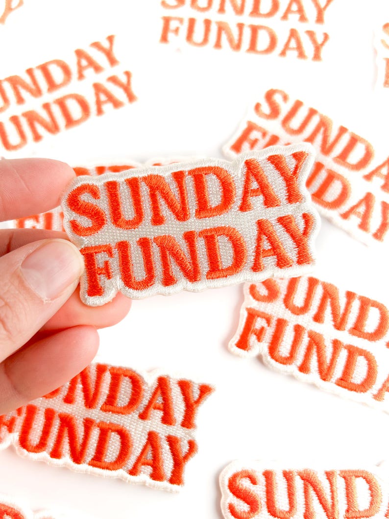 Puede incluir: Parches bordados con las palabras "SUNDAY FUNDAY" en hilo naranja sobre un fondo blanco. Los parches son rectangulares y tienen un borde cosido. Varios parches est&aacute;n dispersos sobre una superficie blanca.