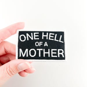 Op de afbeelding: Zwarte stoffen patch met witte tekst die "One Hell of a Mother" luidt.