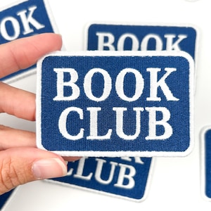 May include: Navy blue rectangular patch with the words "BOOK CLUB" in white, block letters. The patch has a white border and a textured appearance. Other similar patches are visible in the background.